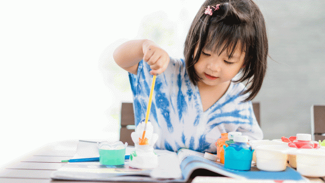 a toddler does arts and crafts with paint and an open book in a blue shirt